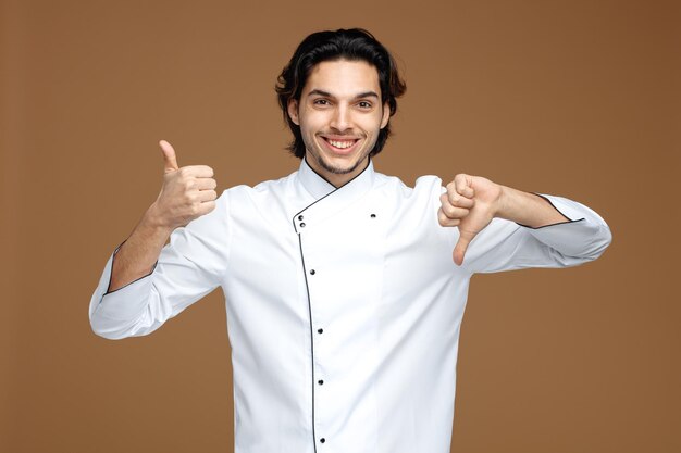uśmiechnięty młody mężczyzna szef kuchni ubrany w mundur, patrzący na kamerę pokazującą kciuki w górę iw dół na białym tle na brązowym tle