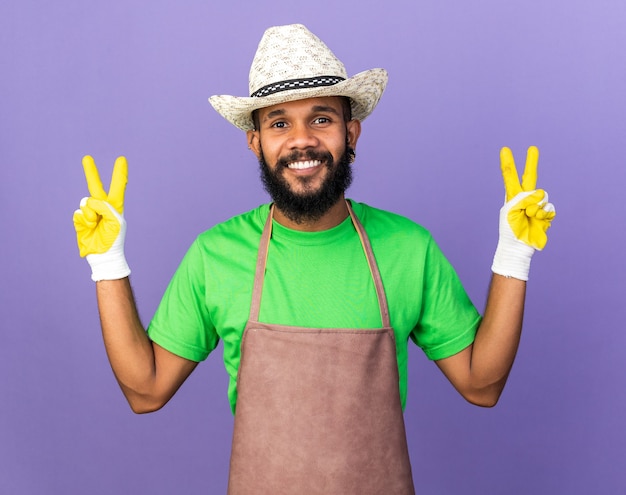 Uśmiechnięty młody ogrodnik afroamerykański facet w kapeluszu ogrodniczym z rękawiczkami pokazującym gest pokoju na niebieskiej ścianie