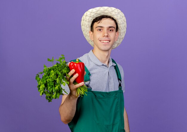 Uśmiechnięty młody ogrodnik męski na sobie kapelusz ogrodniczy posiada czerwoną paprykę i kolendrę