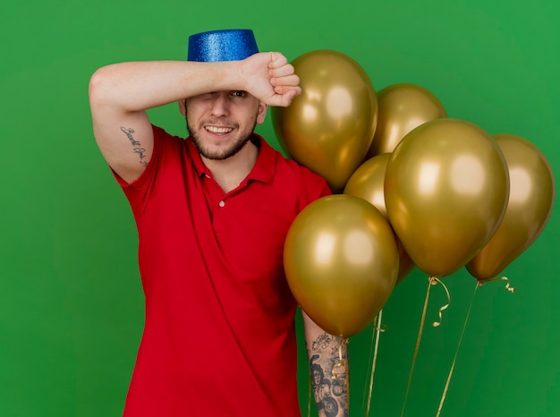 Uśmiechnięty młody przystojny facet słowiańskich partii na sobie kapelusz partii trzymając balony, trzymając ramię przed czołem patrząc na kamery na białym tle na zielonym tle