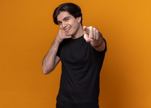 Uśmiechnięty młody przystojny facet ubrany w czarną koszulkę pokazującą rozmowę telefoniczną i punkty w aparacie na białym tle na pomarańczowej ścianie