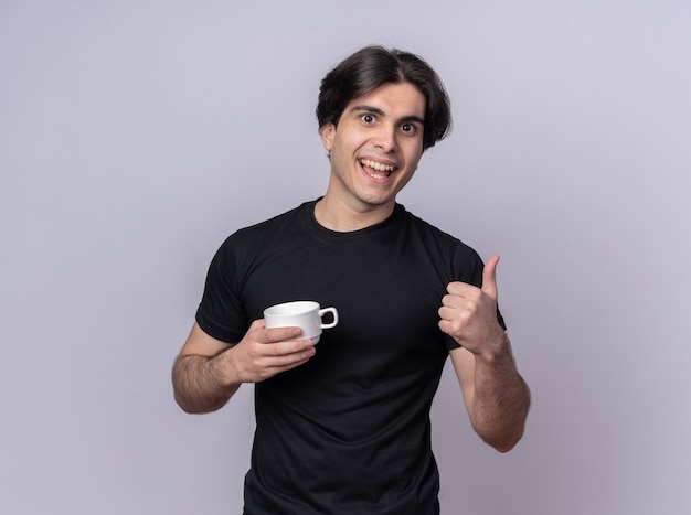 Uśmiechnięty młody przystojny facet ubrany w czarną koszulkę trzymając kubek kawy pokazując kciuk do góry na białym tle na białej ścianie