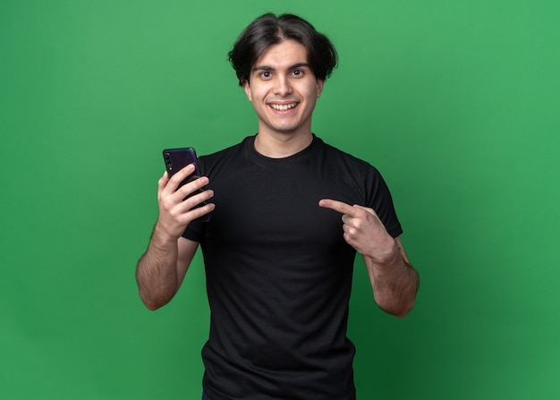 Uśmiechnięty młody przystojny facet ubrany w czarną koszulkę, trzymający i wskazujący na telefon odizolowany na zielonej ścianie