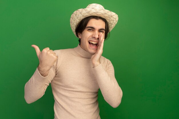 Uśmiechnięty młody przystojny facet w kapeluszu dzwoniąc do kogoś pokazując kciuk do góry na białym tle na zielonej ścianie