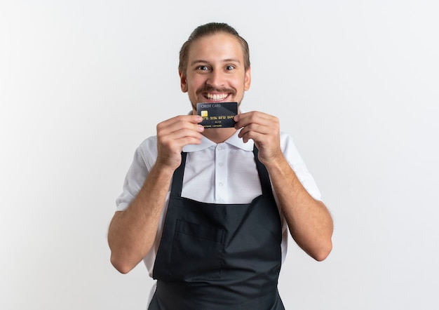 Bezpłatne zdjęcie uśmiechnięty młody przystojny fryzjer na sobie mundur pokazuje kartę kredytową na białym tle na białej ścianie