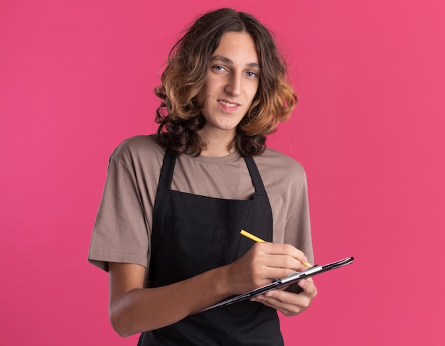 Bezpłatne zdjęcie uśmiechnięty młody przystojny fryzjer ubrany w jednolite pisanie ołówkiem w schowku