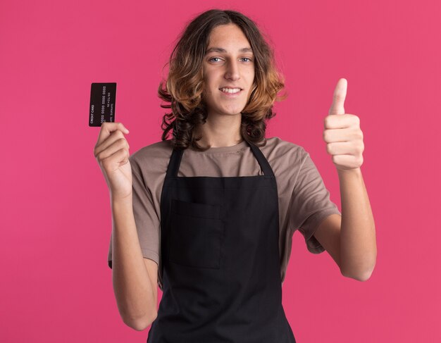 Uśmiechnięty młody przystojny fryzjer ubrany w mundur pokazujący kartę kredytową i kciuk w górę