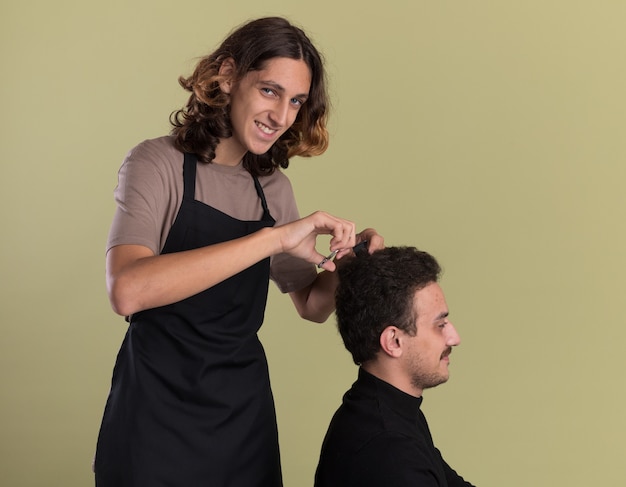 Uśmiechnięty młody przystojny fryzjer w mundurze robi fryzurę dla swojego młodego klienta