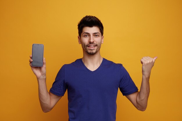 Uśmiechnięty młody przystojny mężczyzna patrzący na kamerę pokazującą telefon komórkowy do kamery wskazującej na bok na żółtym tle