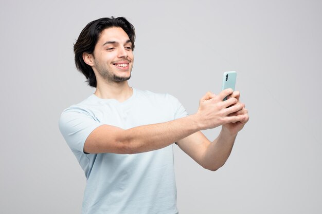 uśmiechnięty młody przystojny mężczyzna rozciągający telefon komórkowy przy selfie na białym tle