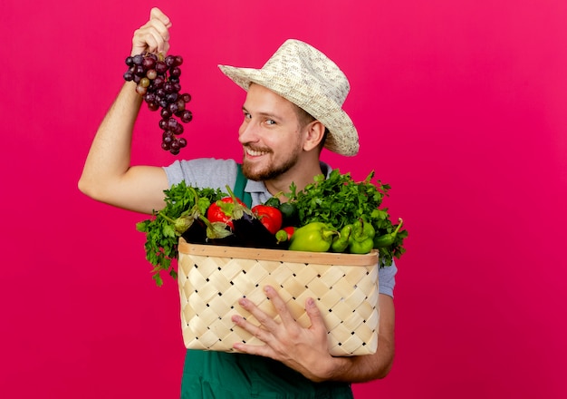 Uśmiechnięty młody przystojny ogrodnik słowiański w mundurze i kapeluszu trzymający kosz warzyw i winogron odizolowany na szkarłatnej ścianie z miejscem na kopię