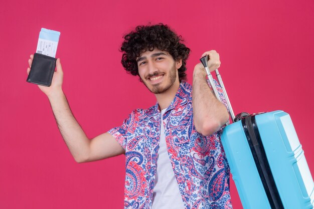 Uśmiechnięty młody przystojny podróżnik kręcone mężczyzna trzyma portfel i bilety lotnicze i walizkę na na białym tle różowej ścianie