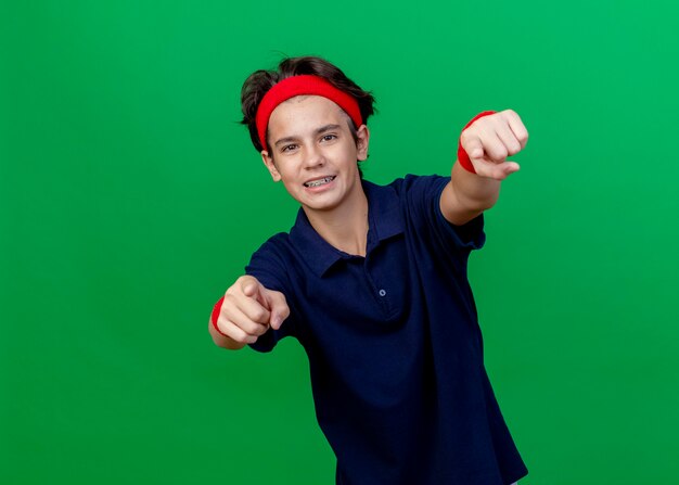 Uśmiechnięty młody przystojny sportowy chłopiec ubrany w opaskę i opaski na nadgarstek z aparatami ortodontycznymi patrząc na aparat, wykonujący gest na białym tle na zielonym tle z miejsca na kopię