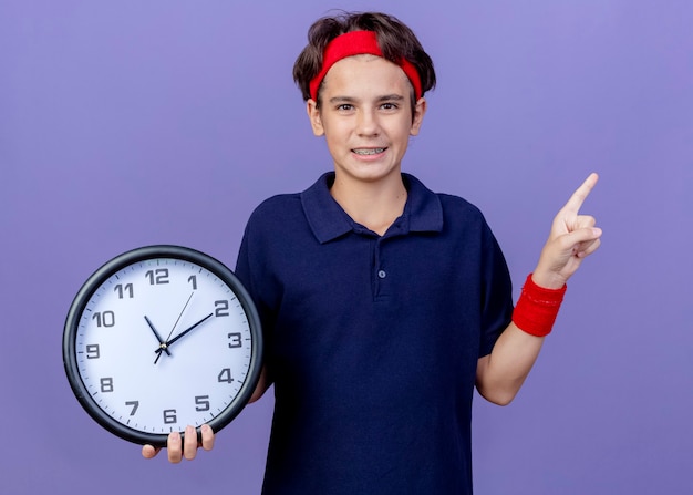 Uśmiechnięty młody przystojny sportowy chłopiec ubrany w opaskę i opaski na rękę z aparatami ortodontycznymi patrząc na aparat trzymający zegar wskazujący na bok na białym tle na fioletowym tle