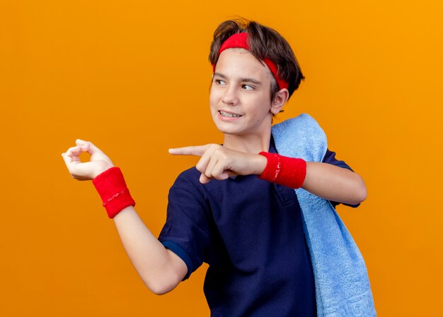 Uśmiechnięty młody przystojny sportowy chłopiec ubrany w opaskę na głowę i opaski na rękę z szelkami dentystycznymi i ręcznikiem na ramieniu patrząc i wskazując na bok na białym tle na pomarańczowym tle