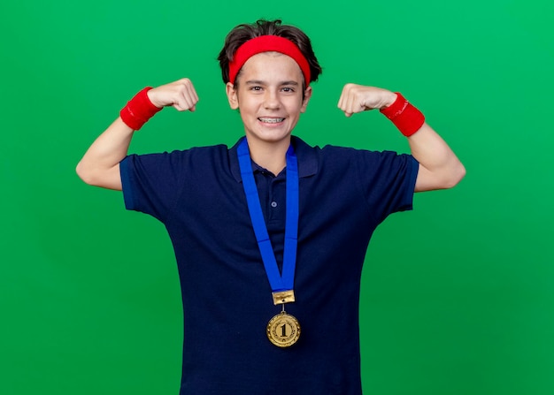 Bezpłatne zdjęcie uśmiechnięty młody przystojny sportowy chłopiec w opasce i opaskach na nadgarstek oraz medal na szyi z aparatami ortodontycznymi patrząc z przodu, robi silny gest odizolowany na zielonej ścianie