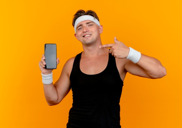Uśmiechnięty młody przystojny sportowy mężczyzna nosi pałąk i opaski na rękę pokazując i wskazując na telefon komórkowy na białym tle na pomarańczowej ścianie