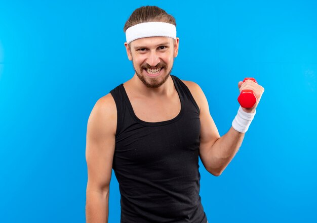Uśmiechnięty młody przystojny sportowy mężczyzna nosi pałąk i opaski na rękę trzymając hantle na białym tle na niebieskiej przestrzeni