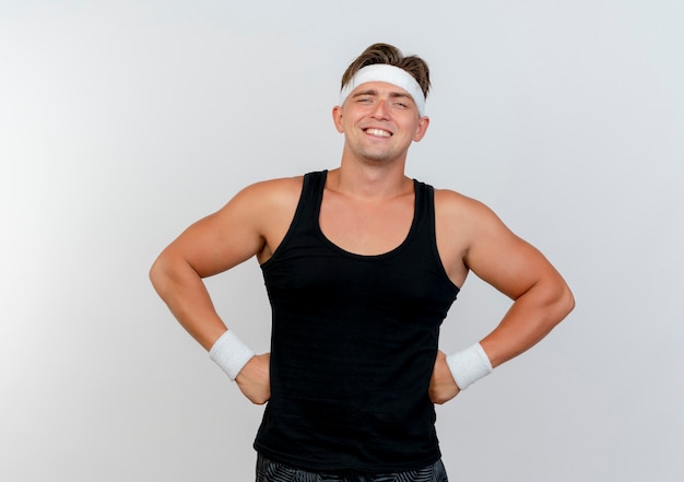 Uśmiechnięty młody przystojny sportowy mężczyzna noszenie opaski i opaski na rękę kładąc ręce na talii na białym tle na białej ścianie