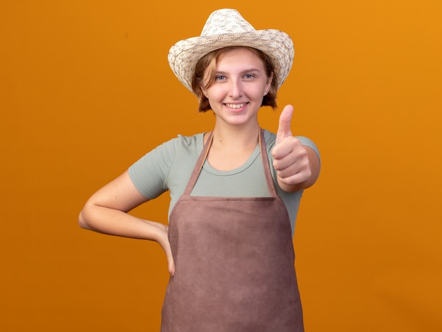 Uśmiechnięty młody słowiański ogrodnik żeński na sobie kapelusz ogrodniczy kciuki do góry na pomarańczowo