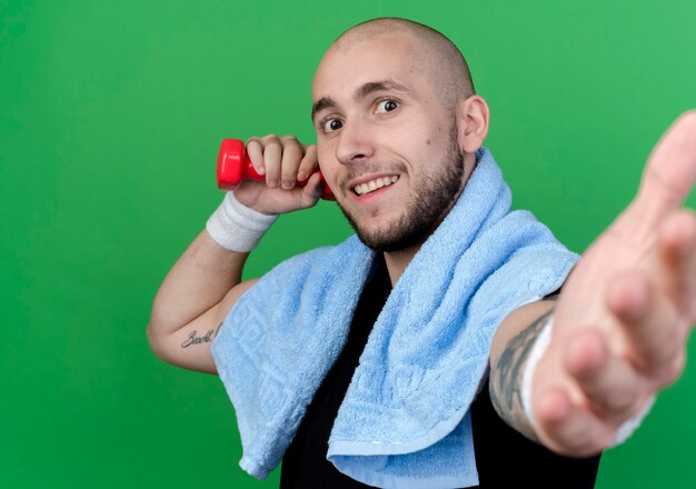 Uśmiechnięty młody sportowy mężczyzna na sobie opaskę, trzymając hantle z ręcznikiem na ramieniu i wyciągając rękę na białym tle na zielonej ścianie