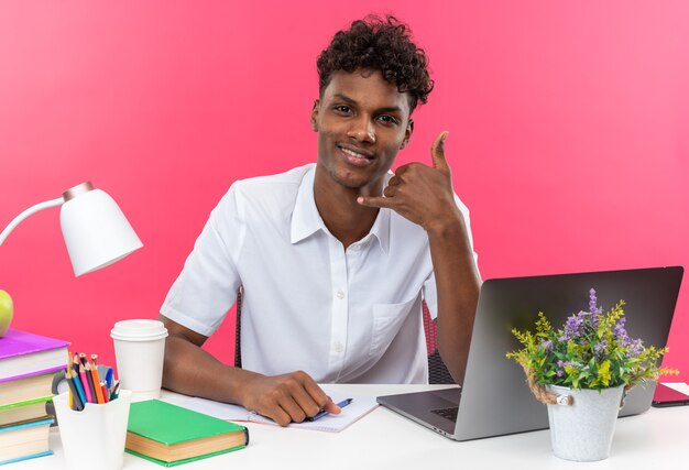 Uśmiechnięty młody student afroamerykański siedzący przy biurku z szkolnymi narzędziami gestykuluje zadzwoń do mnie znak