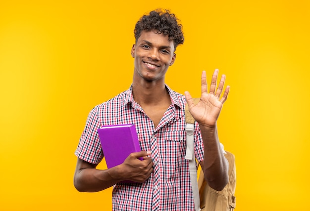 Uśmiechnięty młody student afroamerykański z plecakiem trzymającym książkę i trzymający rękę otwartą na białym tle na pomarańczowej ścianie z miejscem na kopię