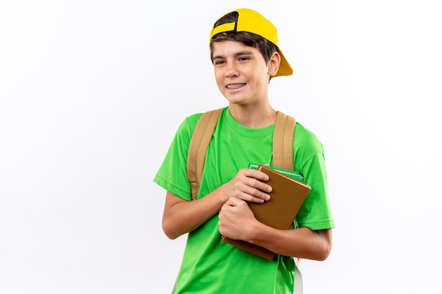 Uśmiechnięty młody szkolny chłopiec ubrany w plecak z czapką trzymający książki