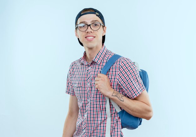 Uśmiechnięty młody uczeń chłopiec jest ubranym tylną torbę i okulary i czapkę na białym tle