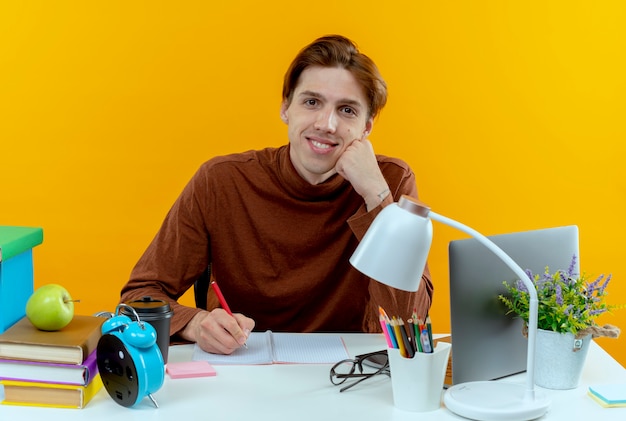 Uśmiechnięty młody uczeń chłopiec siedzi przy biurku z narzędzi szkolnych, pisząc coś na notebooku na żółto