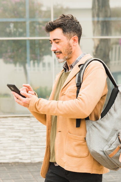 Uśmiechnięty nowożytny mężczyzna z jego plecakiem używać telefon komórkowego