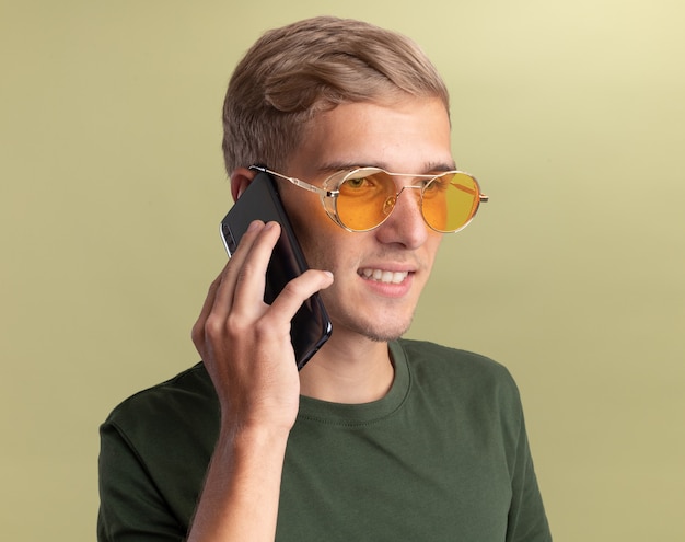Uśmiechnięty, patrząc na bok młody przystojny facet ubrany w zieloną koszulę w okularach mówi przez telefon na białym tle na oliwkowej ścianie