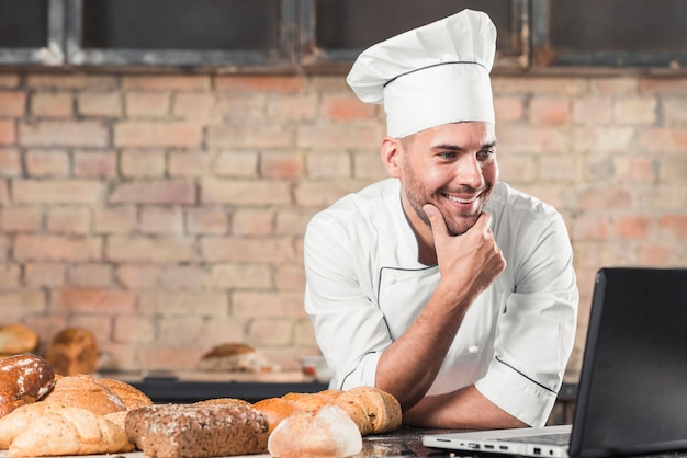 Bezpłatne zdjęcie uśmiechnięty piekarz patrzeje laptop nad kuchennym worktop z piec chlebami