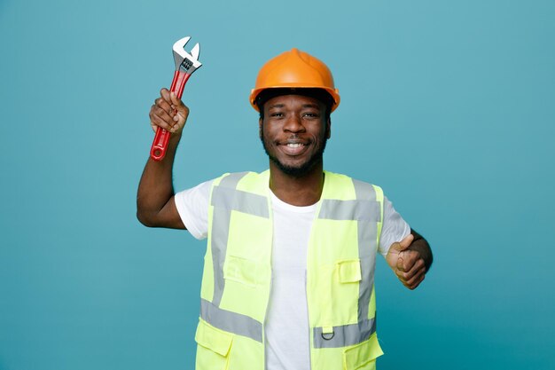 Uśmiechnięty pokazując kciuk do góry młody afroamerykański budowniczy w mundurze trzymający klucz gazowy na białym tle na niebieskim tle