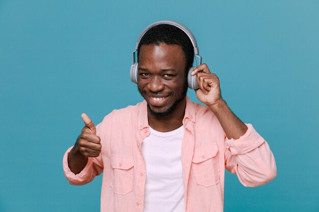 Uśmiechnięty pokazując kciuk do góry młody afroamerykański facet noszący słuchawki na białym tle na niebieskim tle