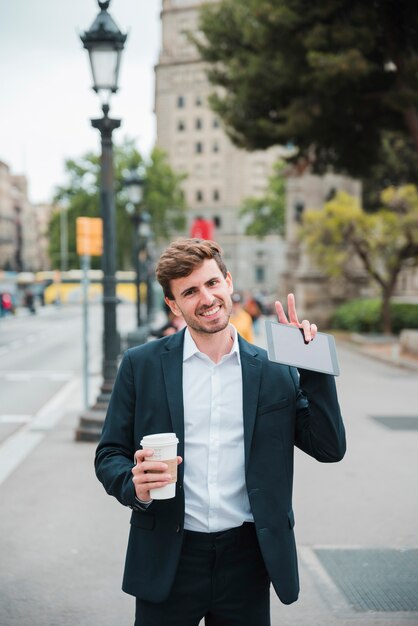Uśmiechnięty portret biznesmen trzyma takeaway filiżankę i cyfrową pastylkę pokazuje zwycięstwo gest