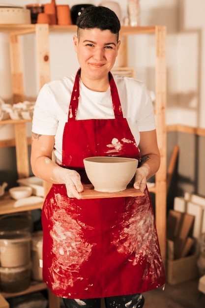 Uśmiechnięty portret młoda kobieta trzyma handmade glinianego puchar na drewnianej tacy w czerwonym fartuchu