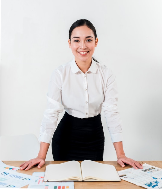 Uśmiechnięty portret młodej bizneswoman stoi za biurkiem przeciw białej ścianie