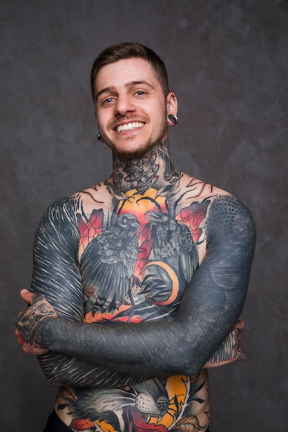 Uśmiechnięty portret tatuażu młody człowiek z przebitym nosem i ucho stoi przeciw popielatemu tłu