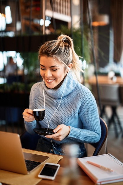 Uśmiechnięty pracownik niezależny pijący kawę i surfujący po sieci na laptopie podczas słuchania muzyki na słuchawkach w kawiarni