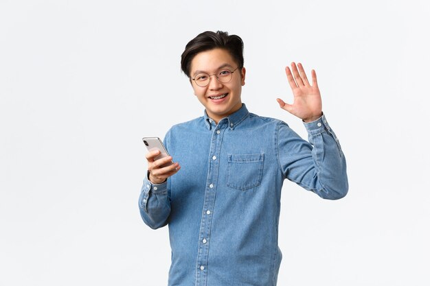 Uśmiechnięty przyjazny azjatycki mężczyzna z szelkami używa telefonu komórkowego, patrząc na kamerę i machając podniesioną ręką, wita się z tobą, znajduje ludzi online w aplikacji randkowej, spotyka się z przyjaciółmi, stoi na białym tle.