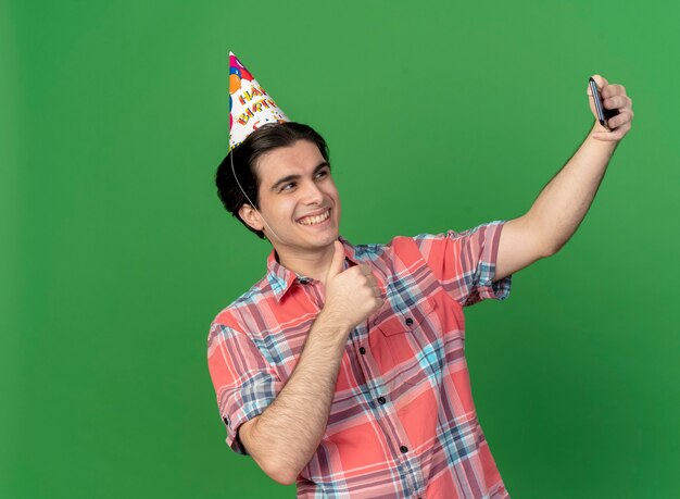 Uśmiechnięty przystojny kaukaski mężczyzna noszący urodzinową czapkę, kciuki w górę, trzymający i patrzący na telefon