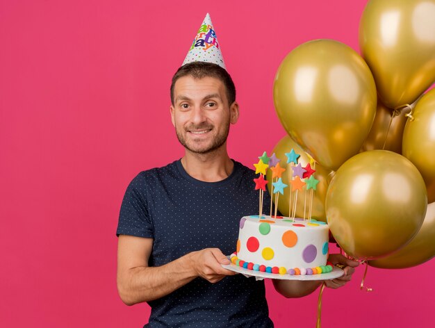 Uśmiechnięty przystojny kaukaski mężczyzna w czapce urodzinowej trzyma tort urodzinowy i balony z helem na białym tle na różowym tle z miejsca na kopię