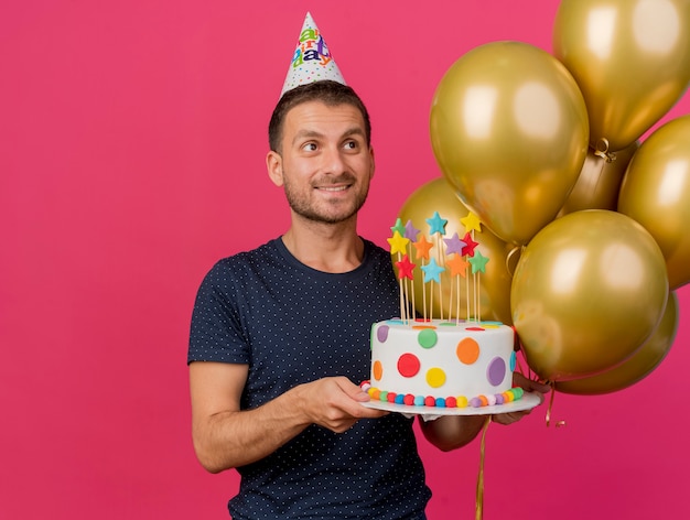 Uśmiechnięty przystojny kaukaski mężczyzna w czapce urodzinowej trzyma tort urodzinowy i balony z helem, patrząc na bok na białym tle na różowym tle z miejsca na kopię