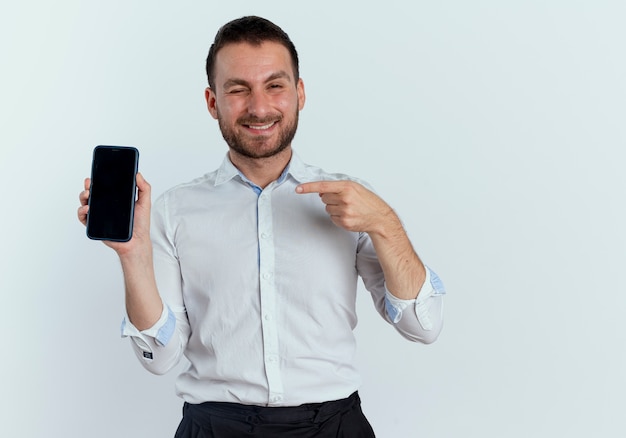 Uśmiechnięty przystojny mężczyzna mruga oczami, trzymając i wskazując na telefon na białym tle na białej ścianie