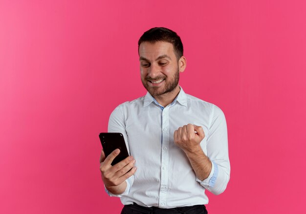 Uśmiechnięty przystojny mężczyzna trzyma pięść trzymając i patrząc na telefon na białym tle na różowej ścianie