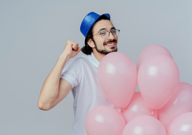 Uśmiechnięty przystojny mężczyzna w okularach i niebieskim kapeluszu trzyma balony i pokazuje tak gest na białym tle