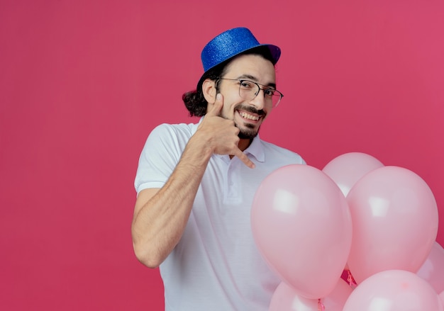 Bezpłatne zdjęcie uśmiechnięty przystojny mężczyzna w okularach i niebieskim kapeluszu trzyma balony i pokazuje telefon gest na białym tle na różowym tle