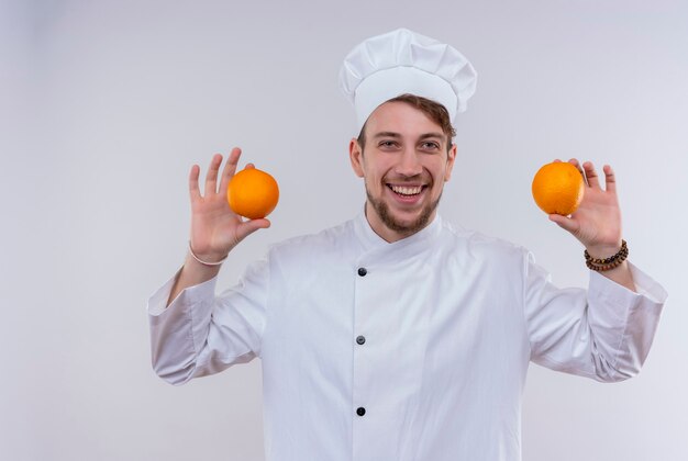 Uśmiechnięty przystojny młody brodaty mężczyzna kucharz ubrany w biały mundur kuchenki i kapelusz gospodarstwa pomarańcze, patrząc na białej ścianie