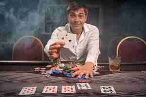 Bezpłatne zdjęcie uśmiechnięty rozradowany facet w białej koszuli gra w pokera, siedząc przy stole w kasynie w dymie. cieszy się z wygranej pokazując dwa asy przed kamerą. hazard za pieniądze. gry losowe.